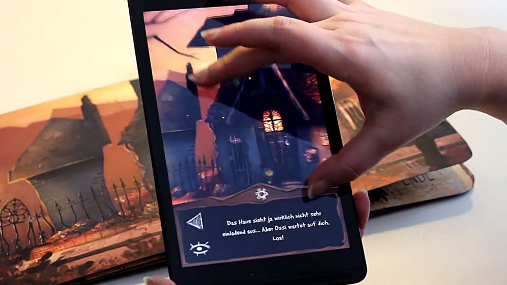Ein Handybildschirm mit der App zu Ernas Unheil ist über dem Buch zum Spiel. Eine Hand bedient die App